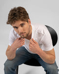 Man wearing a white t-shirt and blue jeans sitting on a black chair against a gray background