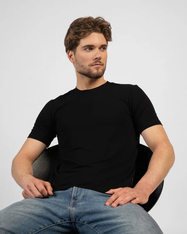 Man wearing a black t-shirt and blue jeans sitting on a chair against a plain background