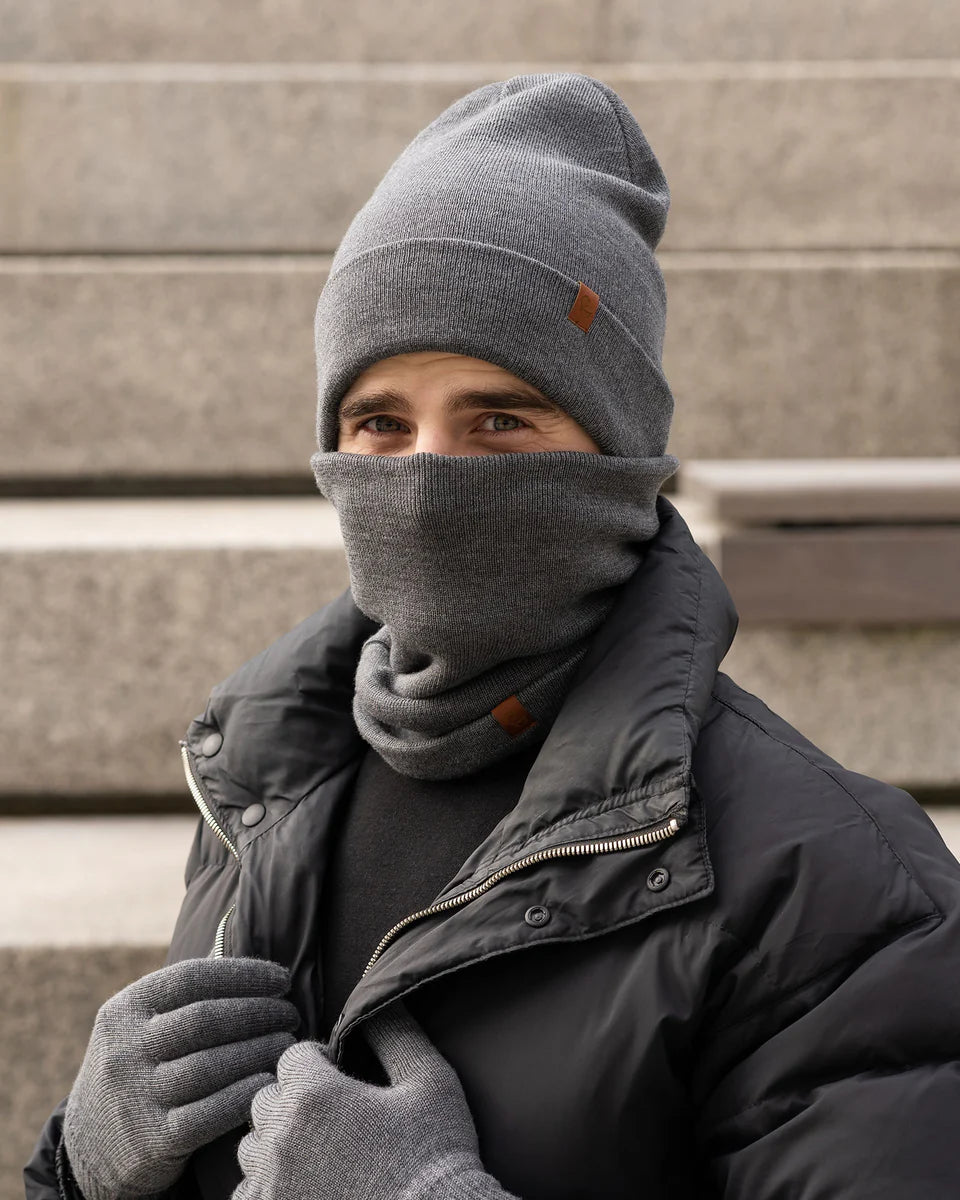 Person wearing a gray beanie, scarf, and gloves in front of a concrete wall.