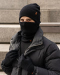 Person wearing a black beanie, scarf, gloves, and coat against a stone wall.