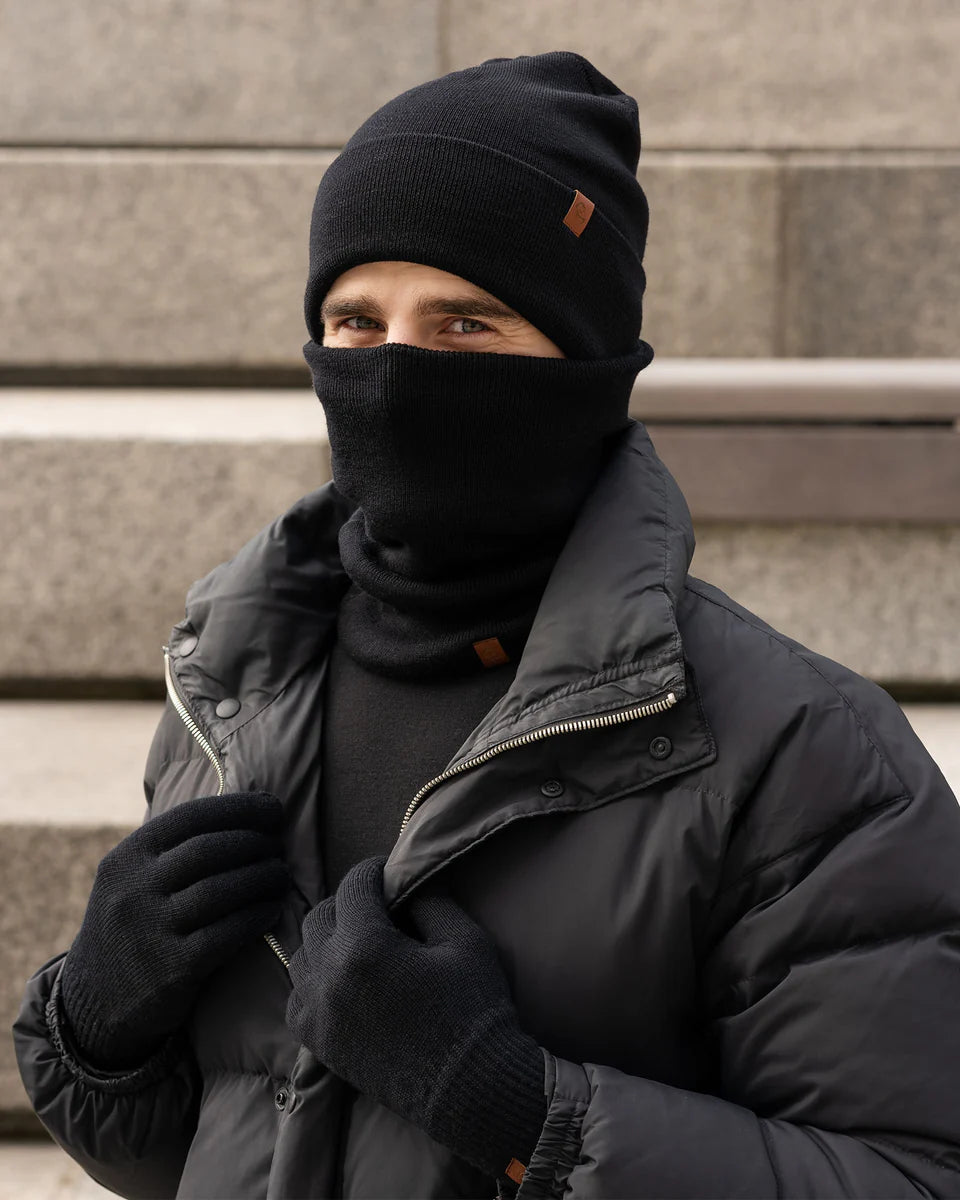 Person wearing a black beanie, scarf, gloves, and coat against a stone wall.