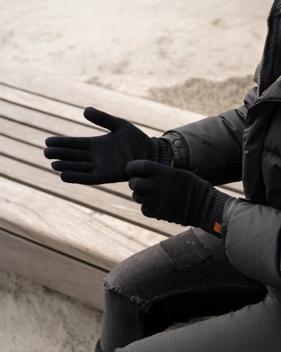 Person wearing black gloves and a dark jacket sitting on a wooden bench.