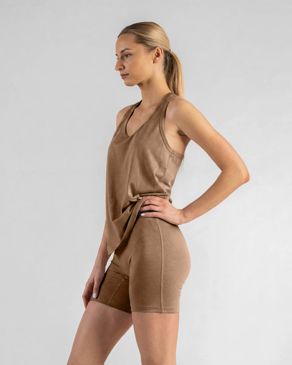 A woman modeling a matching tan activewear set consisting of a sleeveless wrap-style tank top and high-waisted fitted shorts, photographed against a light gray studio background.