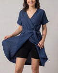 A woman wearing a navy blue linen wrap dress with short sleeves and a tie waist, styled over black bike shorts, against a plain light gray background.