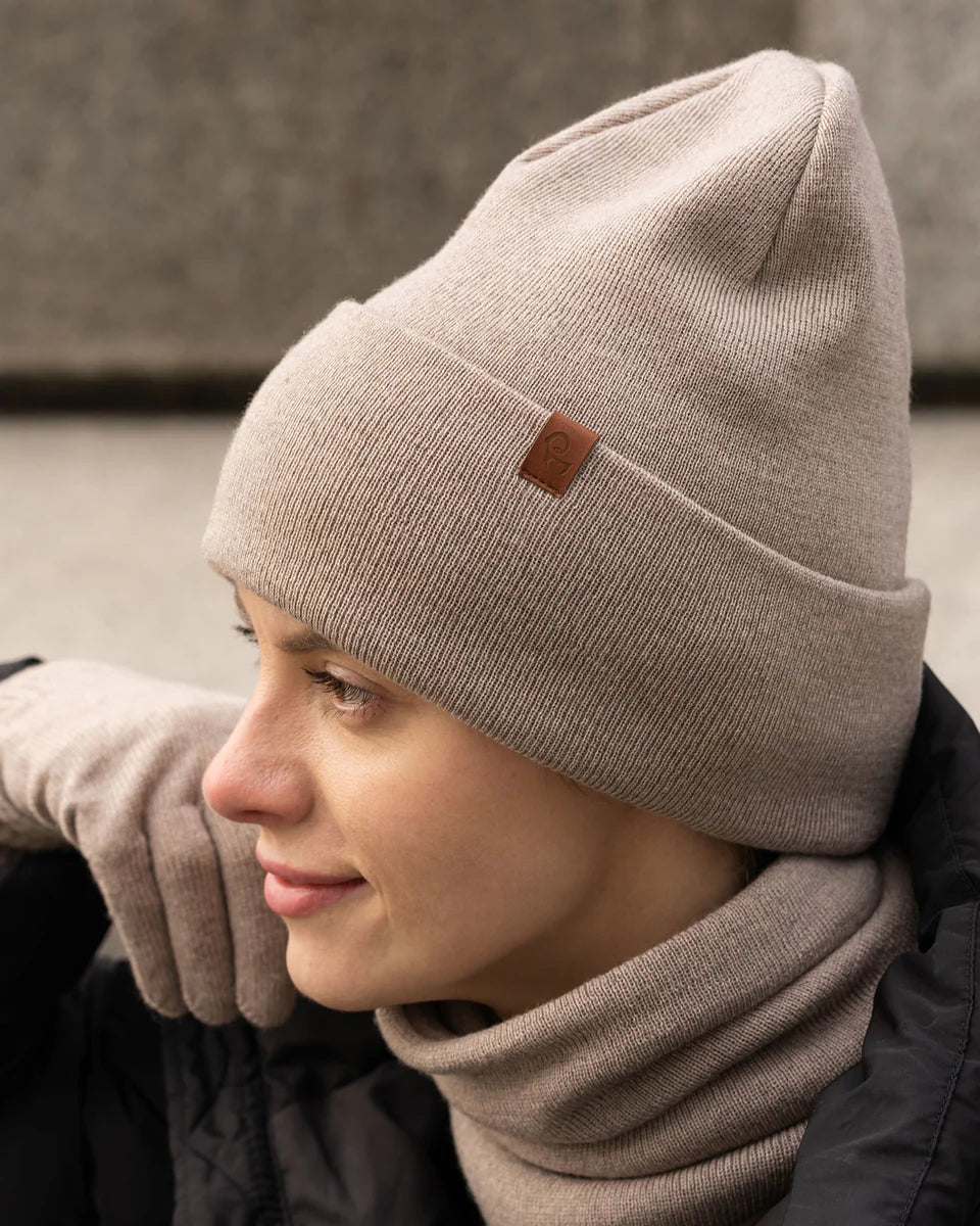 Person wearing a beige beanie and scarf with a brand logo, standing against a neutral background.