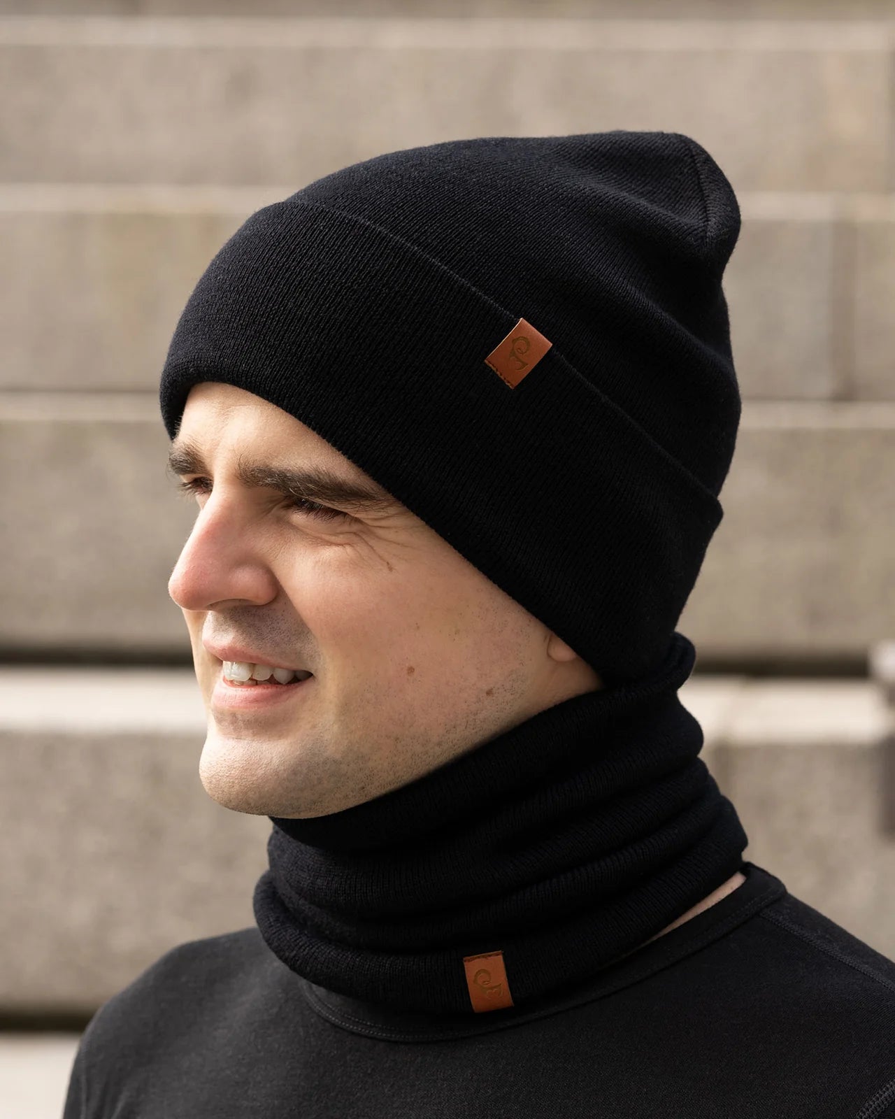 Man wearing a black knit beanie and scarf with brown accents against a stone wall background