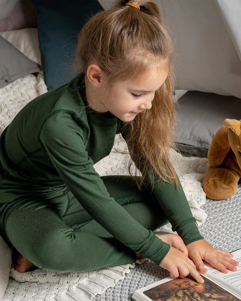 Young girl in a green outfit sitting on a blanket, reading a book with a brown teddy bear beside her.