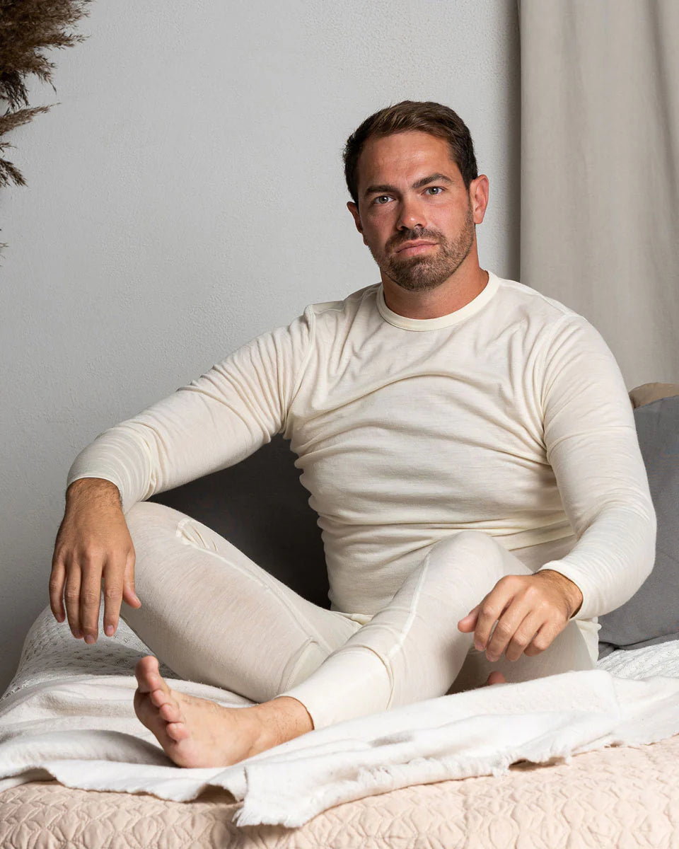 Man wearing a white long-sleeve shirt and pants sitting on a bed.
